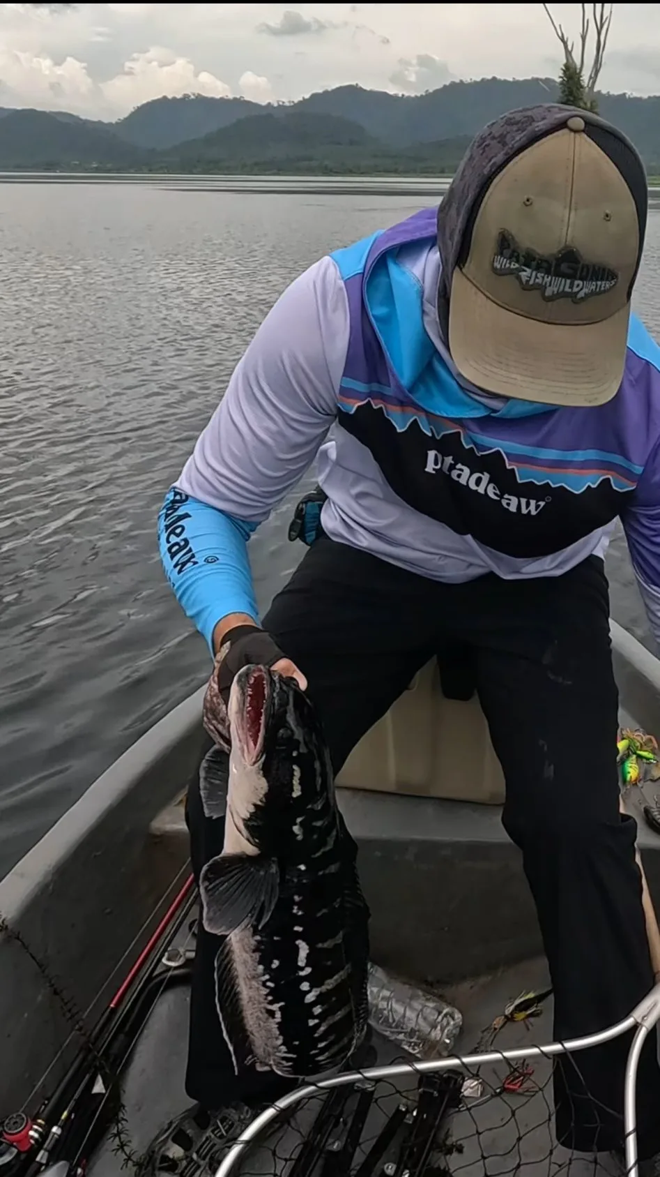 Angler Joshua Rittmann holding a large giant snakehead by the jaw in a fishing boat on a Chanthaburi reservoir, with lures and rods visible in the background.