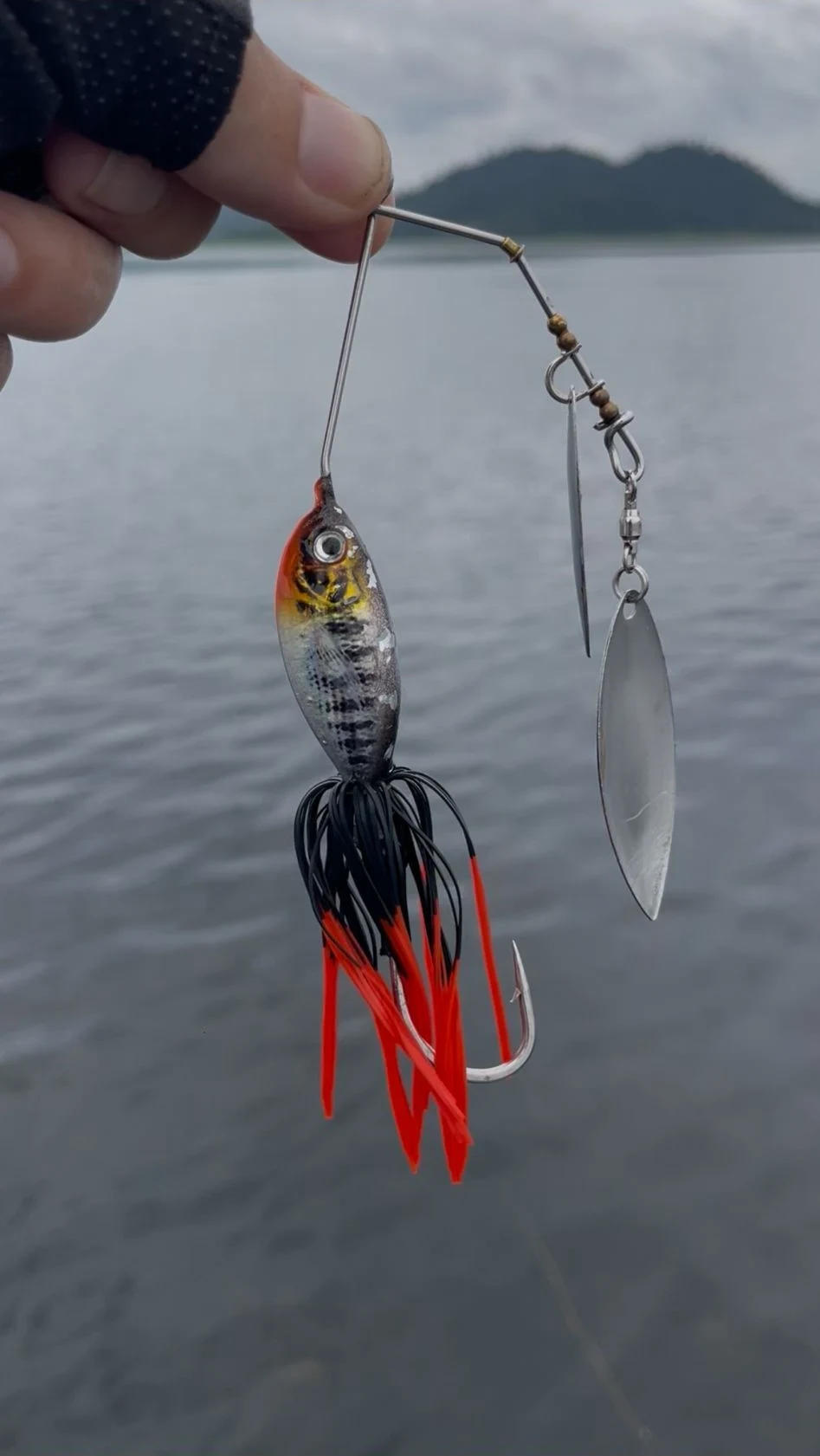 Hand holding a double-bladed spinnerbait with a black and red rubber skirt and fish-shaped body against a Chanthaburi reservoir and mountain backdrop in Thailand.