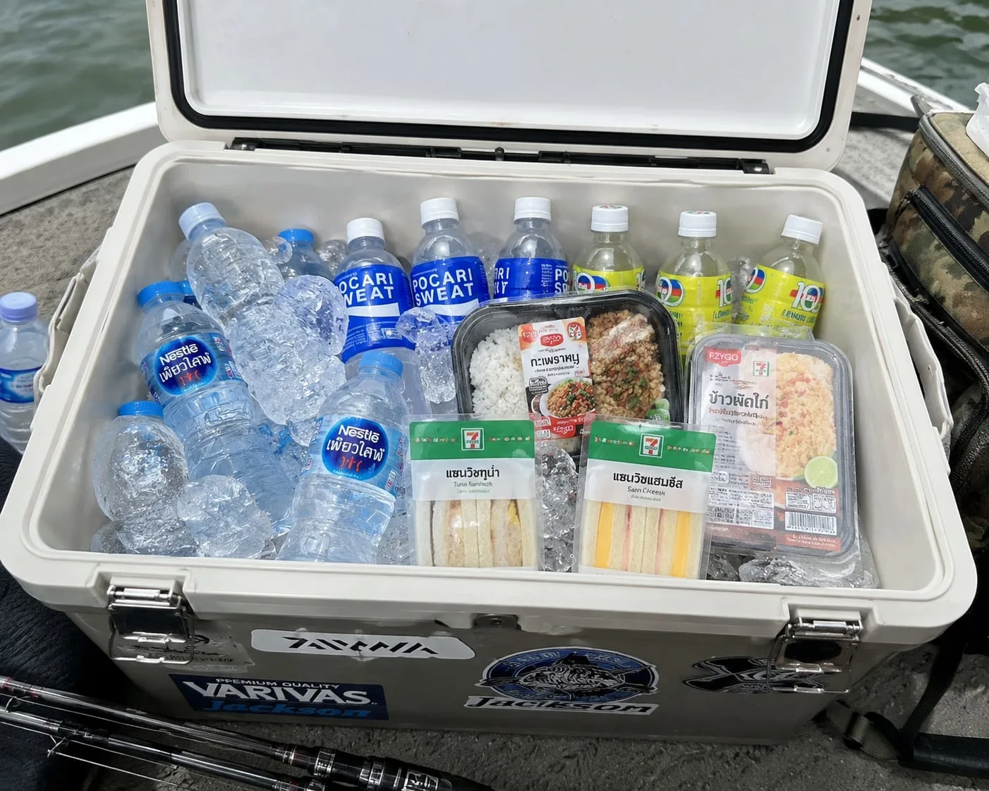 Daiwa cooler box packed with Nestlé water, Pocari Sweat electrolyte drinks, 7-Eleven tuna and ham cheese sandwiches, and Thai rice meals, sitting on a fishing boat in Thailand.