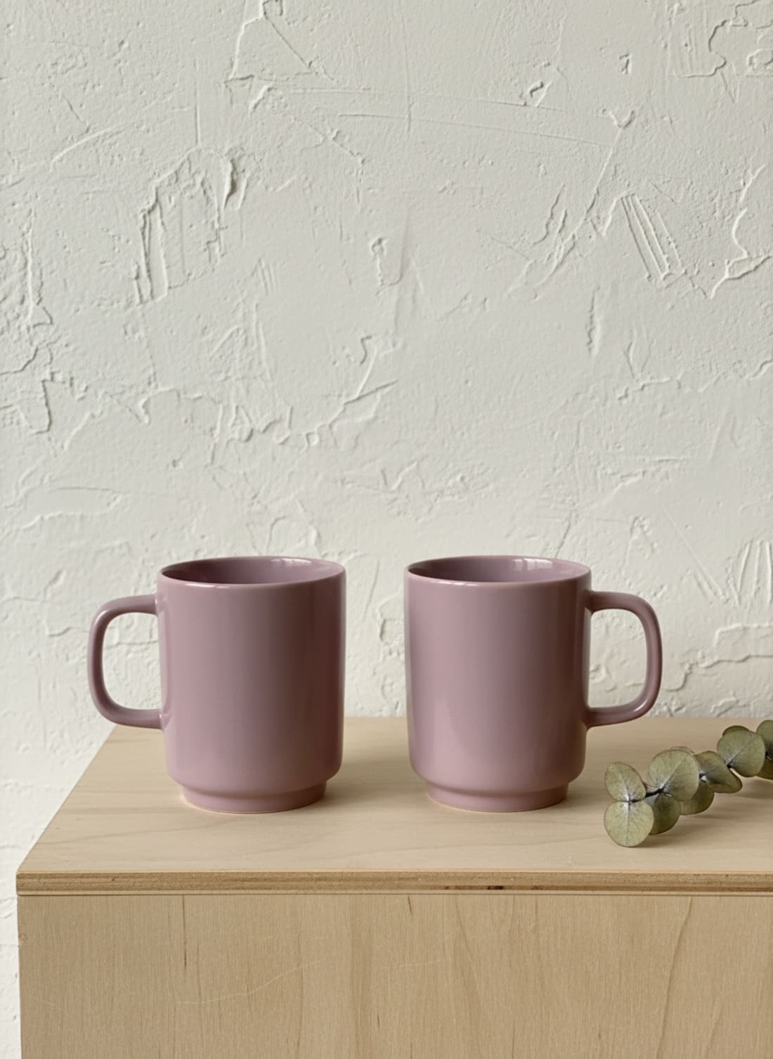 A set of two minimalist dusty rose or mauve ceramic mugs with a glossy finish, placed on a light wood surface with a textured background.
