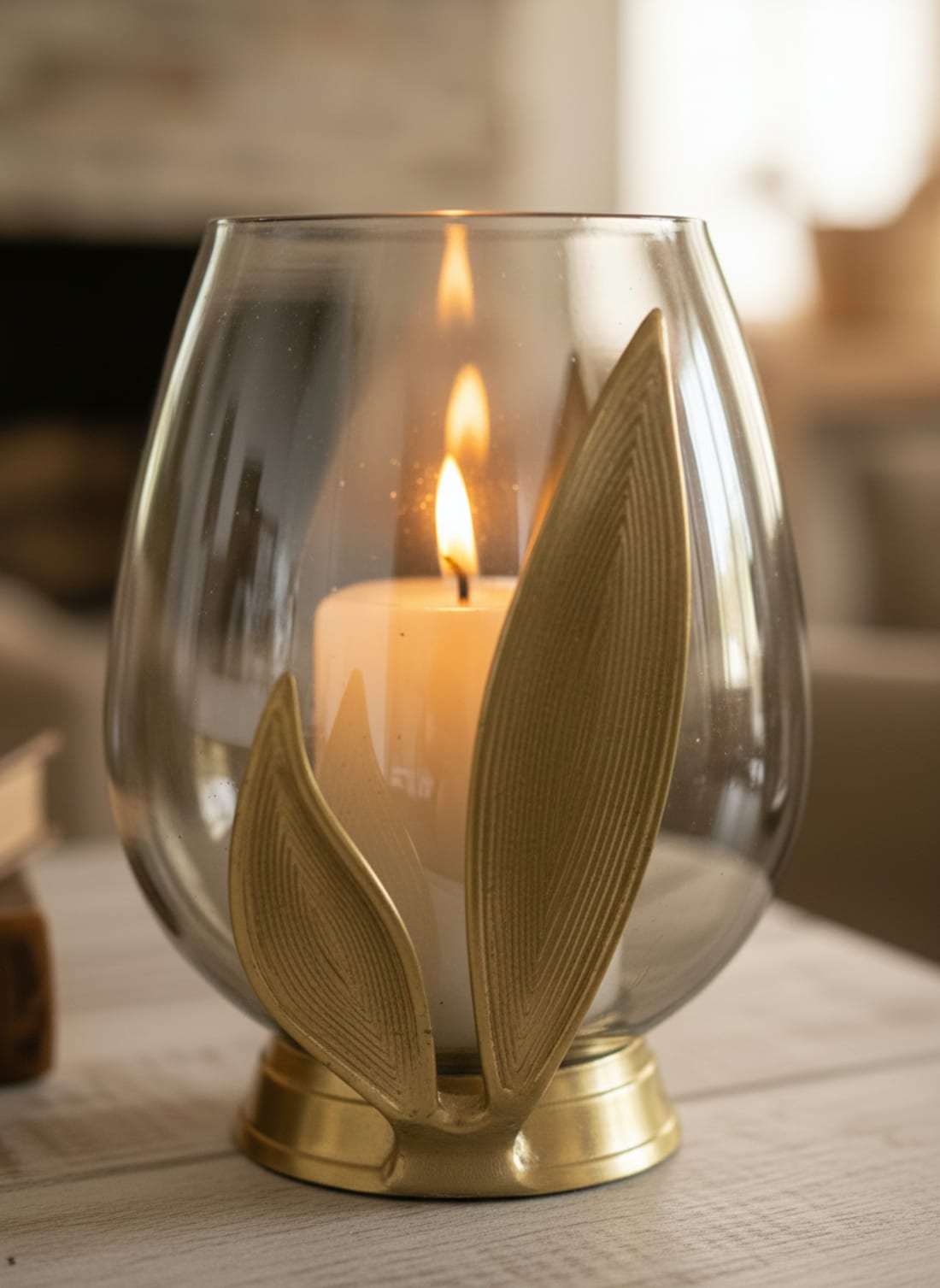 Modern clear glass hurricane candle holder with a decorative golden brass leaf base featuring a lit pillar candle.