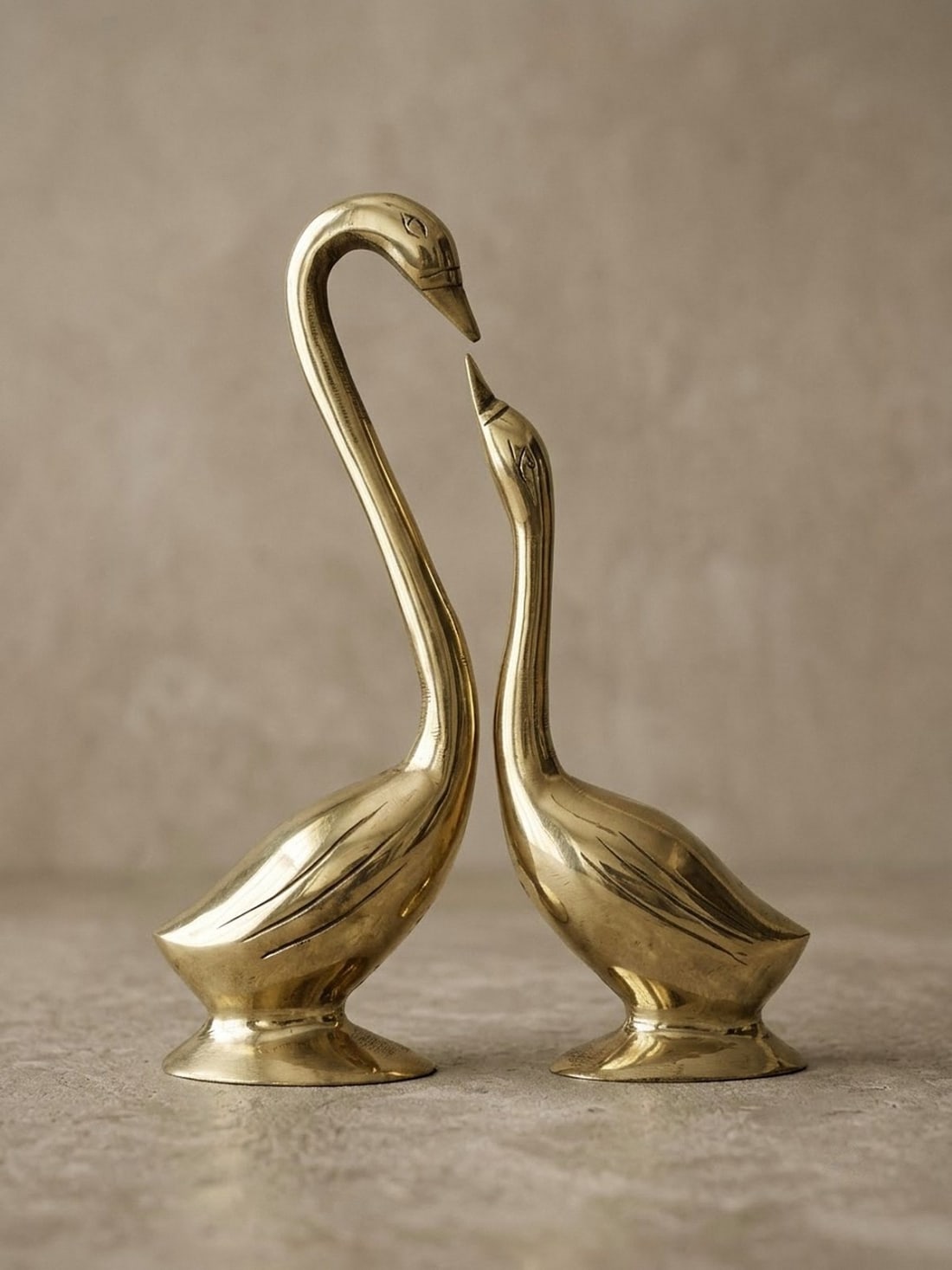 Pair of polished brass swans on oval pedestals facing each other, the taller swan's bowed neck and the smaller swan's raised beak forming a heart