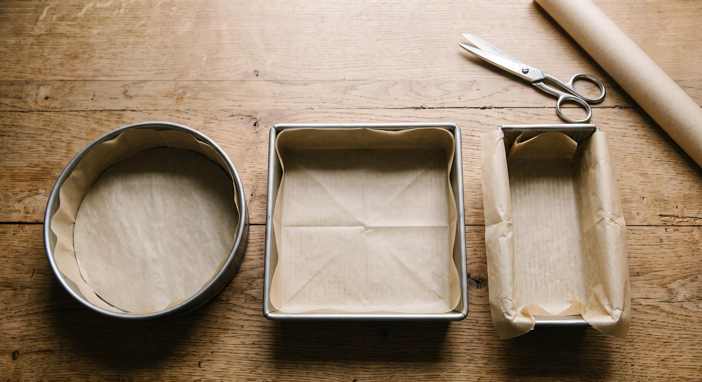 Round, square and loaf cake tins neatly lined with baking paper on a wooden worktop