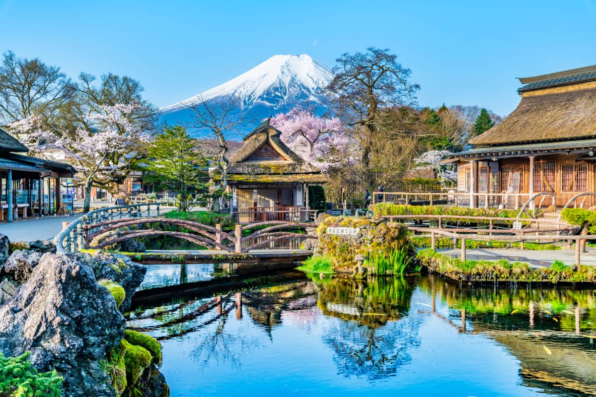 Crystal-clear spring ponds of Oshino Hakkai surrounded by traditional thatched-roof houses, walking paths, and rural village scenery with Mount Fuji rising in the background under a clear sky in Yamanashi, Japan.