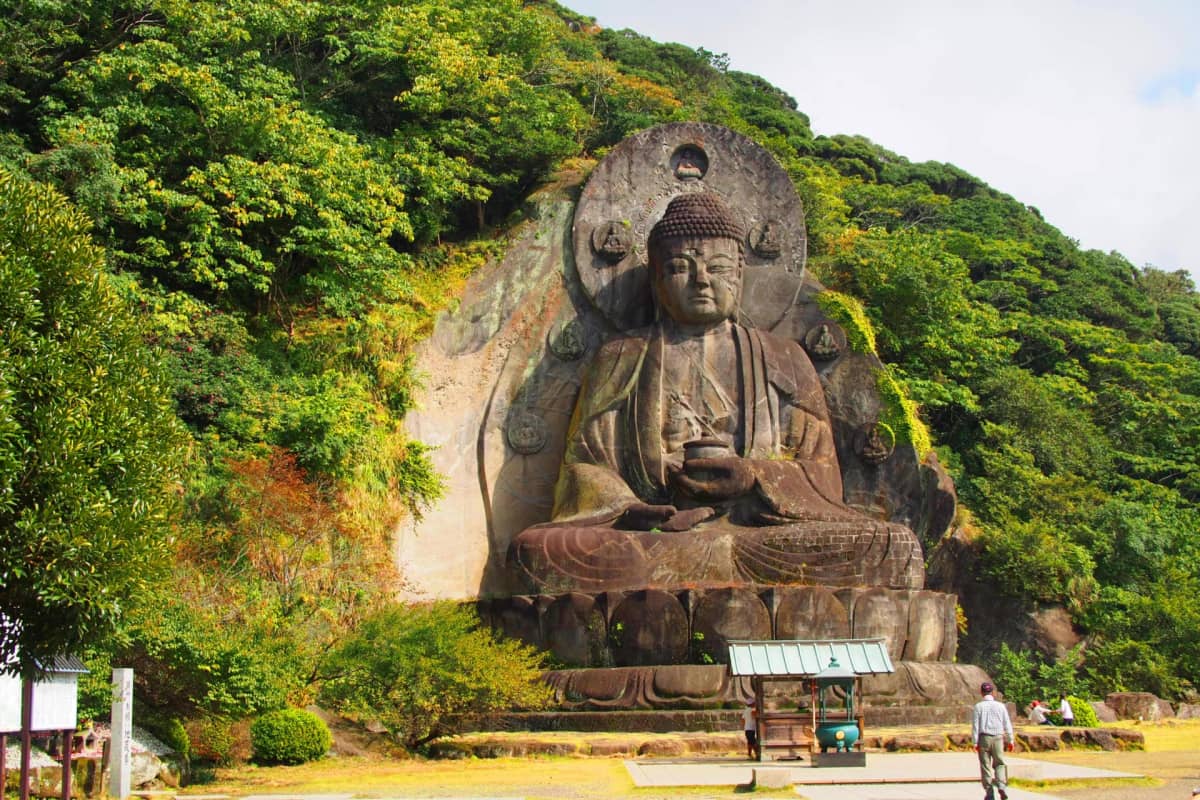 Nihon-ji Daibutsu seated against a rugged rock cliff with lush greenery in the sacred grounds of Nihon-ji Temple.