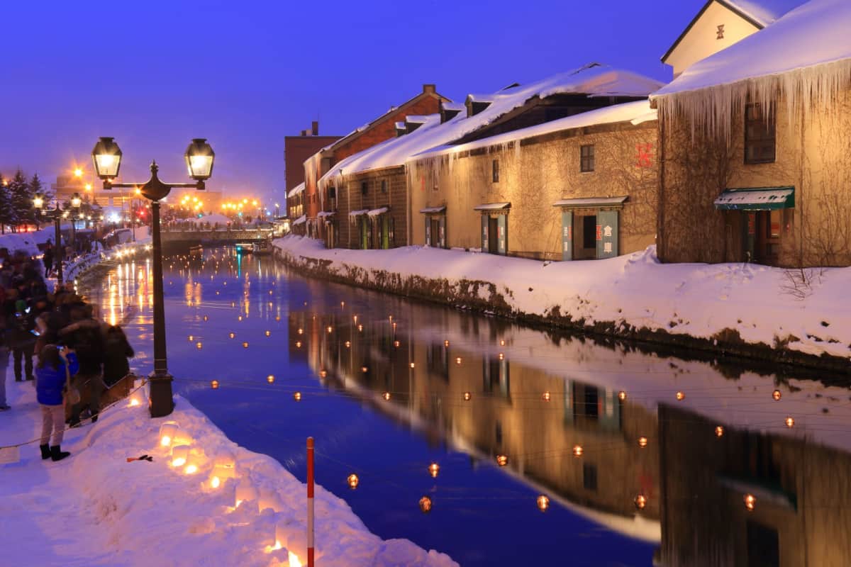 The iconic Otaru Canal lined with historic warehouses, vintage gas lamps, and reflections shimmering on the water at sunset.