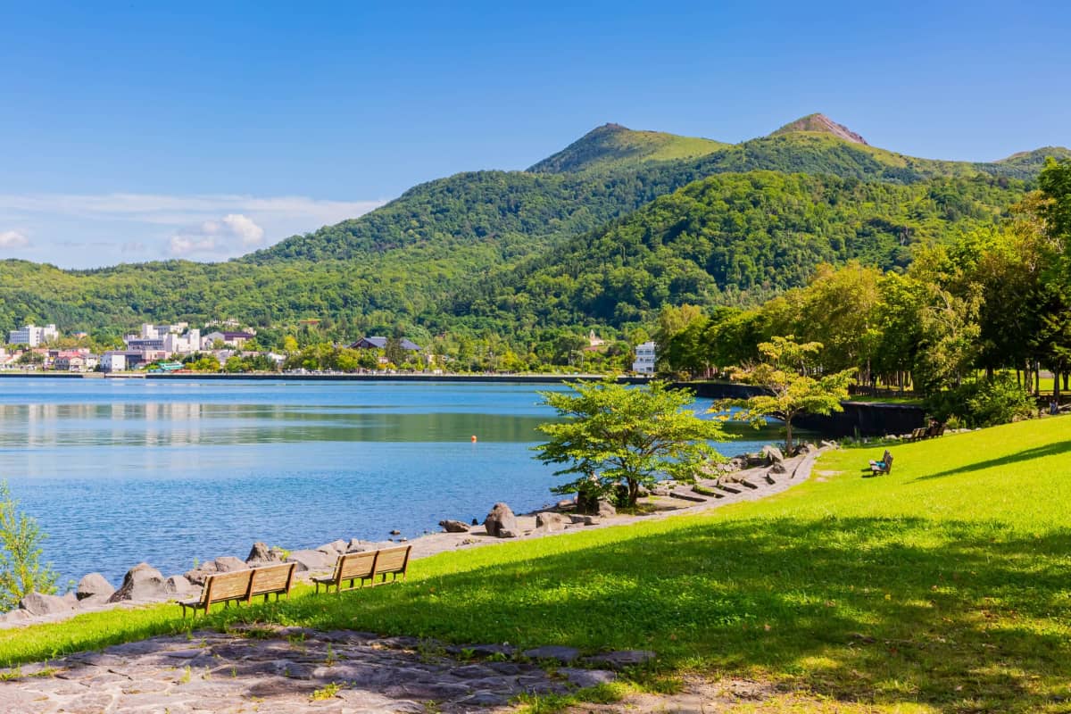 Scenic view of Lake Toya in Hokkaido, with calm waters reflecting the surrounding mountains, forested islands, lakeside hot spring town, and the natural landscapes of Shikotsu-Toya National Park.