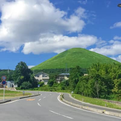 A scenic view of Mount Omuro’s perfectly shaped volcanic crater in Izu, with visitors walking along the circular ridge trail, overlooking sweeping views of Mount Fuji, Sagami Bay, and the surrounding green landscape under open skies.