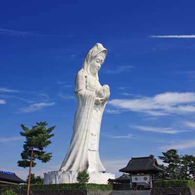 A towering pale pink Kannon statue rises above peaceful Japanese gardens in Aizuwakamatsu, surrounded by koi ponds, pagodas, seasonal trees, and mountain views under the open Fukushima sky.