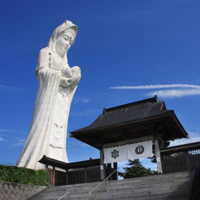 A towering pale pink Kannon statue rises above peaceful Japanese gardens in Aizuwakamatsu, surrounded by koi ponds, pagodas, seasonal trees, and mountain views under the open Fukushima sky.