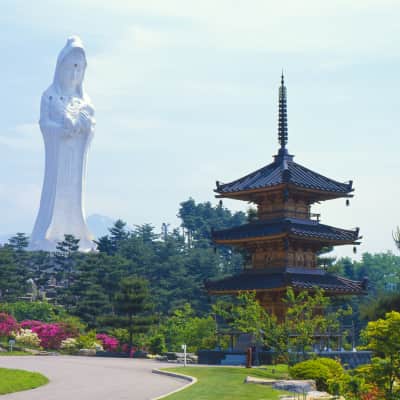 A towering pale pink Kannon statue rises above peaceful Japanese gardens in Aizuwakamatsu, surrounded by koi ponds, pagodas, seasonal trees, and mountain views under the open Fukushima sky.