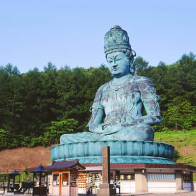 The towering Showa Daibutsu bronze Buddha statue rises above the peaceful grounds of Seiryuji Temple in Aomori, surrounded by trees and traditional Buddhist architecture under a clear blue sky.