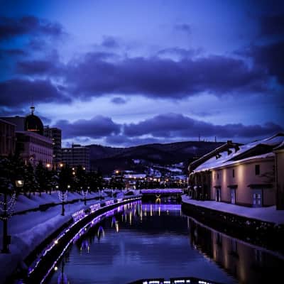 Historic stone warehouses and glowing gas lamps reflected in the calm waters of Otaru Canal during blue hour in Hokkaido, Japan.