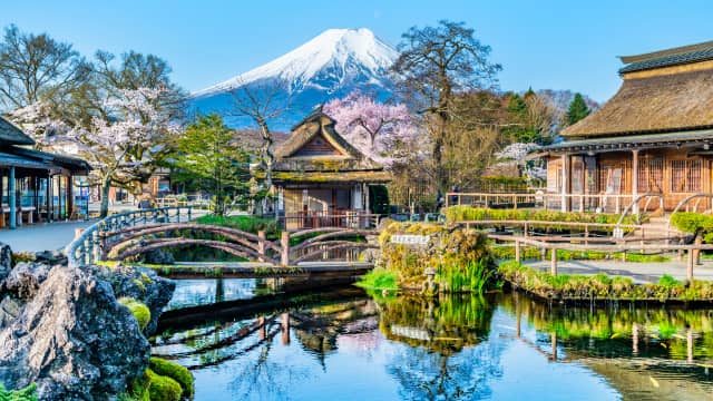 Crystal-clear spring ponds of Oshino Hakkai surrounded by traditional thatched-roof houses, walking paths, and rural village scenery with Mount Fuji rising in the background under a clear sky in Yamanashi, Japan.