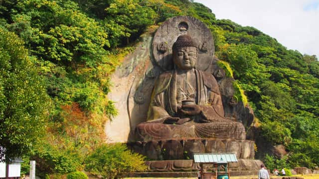 Nihon-ji Daibutsu seated against a rugged rock cliff with lush greenery in the sacred grounds of Nihon-ji Temple.