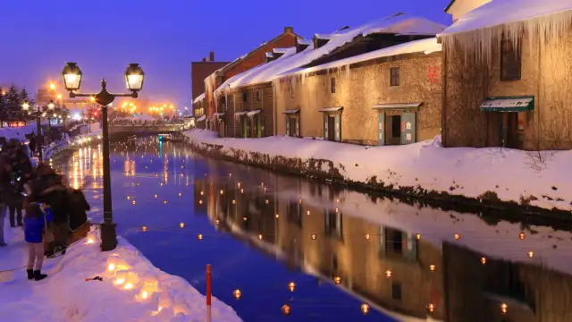 The iconic Otaru Canal lined with historic warehouses, vintage gas lamps, and reflections shimmering on the water at sunset.