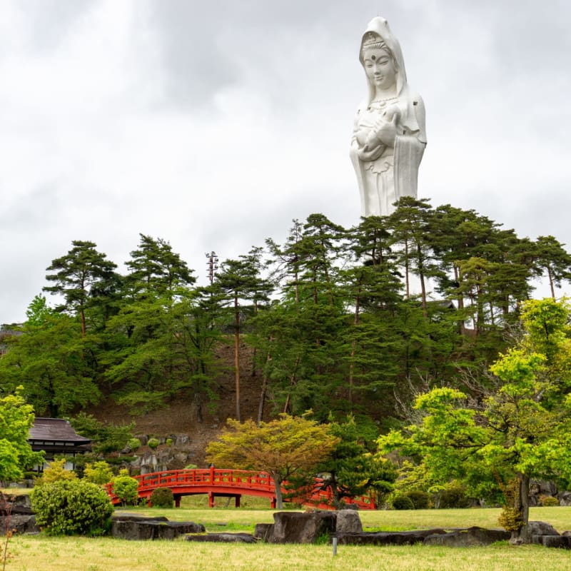A towering pale pink Kannon statue rises above peaceful Japanese gardens in Aizuwakamatsu, surrounded by koi ponds, pagodas, seasonal trees, and mountain views under the open Fukushima sky.