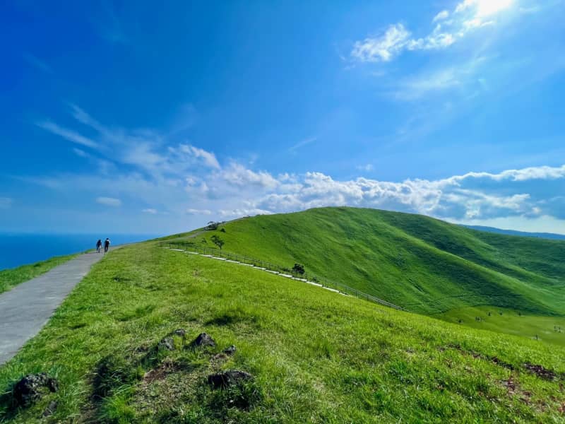 A wide, elevated view of Mount Omuro’s perfectly circular crater, with the green ridge trail wrapping around the summit and distant views stretching toward Mount Fuji and the coastline under a clear sky.