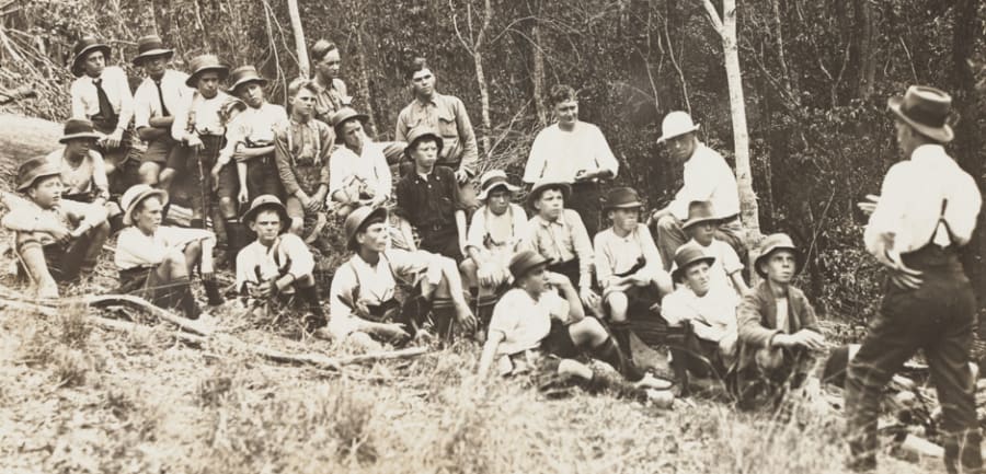 A black and white photograph showing the back of a man wearing a hat, turned towards a group of adolescent boys. They are outdoors on dry grass in front of trees.