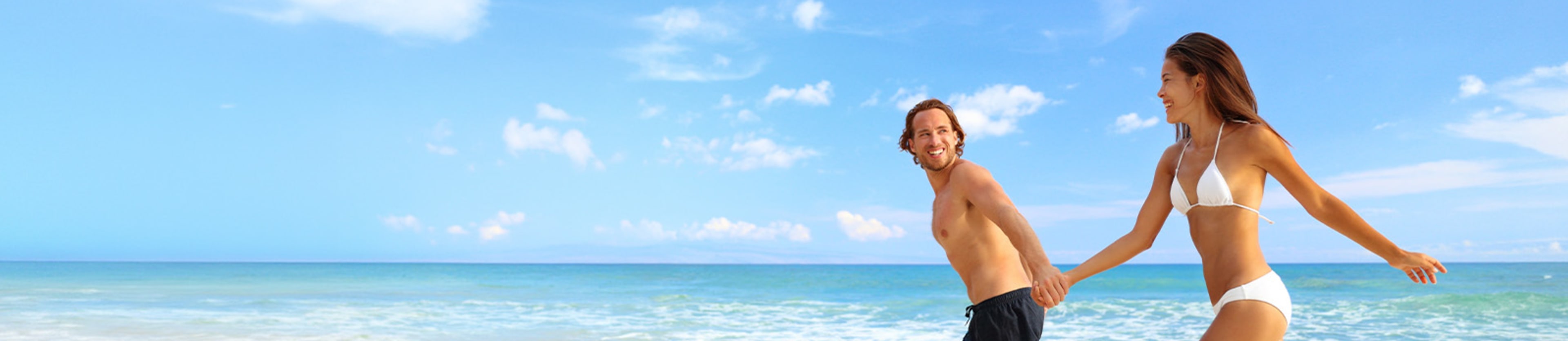 Two people in swimwear walking along the beach, with the ocean and a clear blue sky in the background.