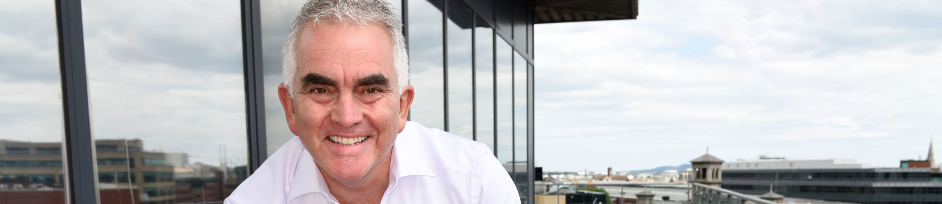 A smiling man in a white shirt sits on a pink chair on a balcony overlooking a cityscape.