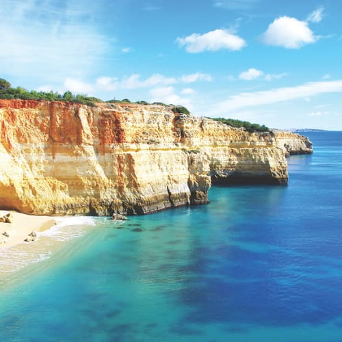 A stunning coastal landscape with towering cliffs, a pristine sandy beach, and crystal-clear turquoise waters against a vibrant blue sky dotted with fluffy white clouds.