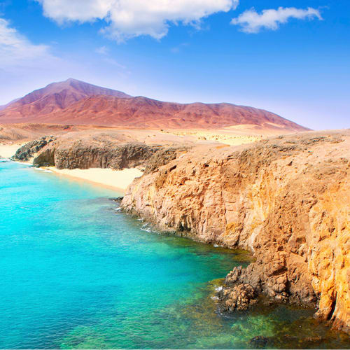 A stunning coastal landscape with rugged cliffs, turquoise waters, and a majestic mountain range in the background under a vibrant blue sky with fluffy clouds.