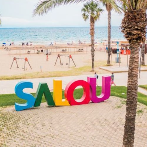Colorful letters spelling "SALOU" are prominently displayed in the foreground, surrounded by palm trees and a beach scene with people in the background.