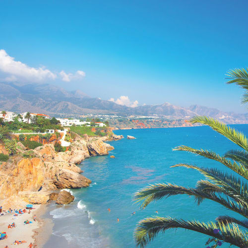 A picturesque coastal scene with a vibrant blue ocean, rocky cliffs, and palm trees in the foreground, set against a backdrop of mountains and a clear blue sky.