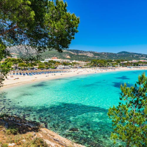 A picturesque beach with crystal-clear turquoise waters, surrounded by lush green trees and a mountainous landscape in the background.