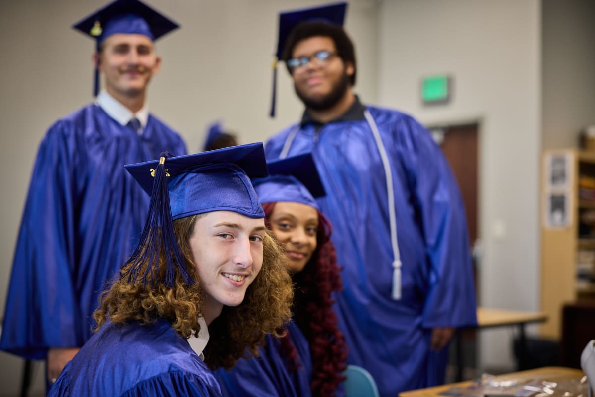 A SailFuture Academy graduate in a blue cap and gown smiling at the camera, with classmates behind