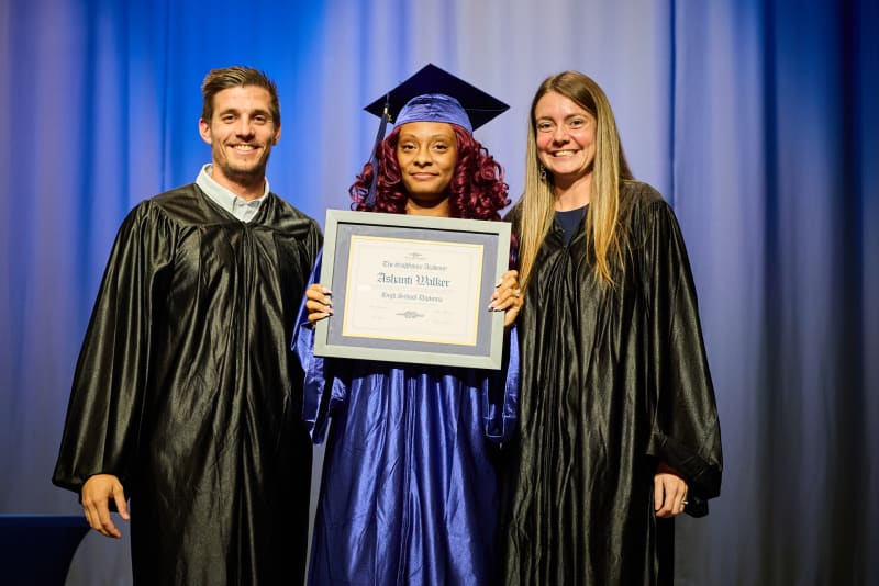 A graduate in cap and gown holding a diploma between two teachers