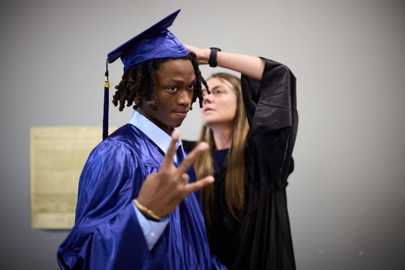 A graduate flashing a peace sign while a teacher straightens their cap before the ceremony