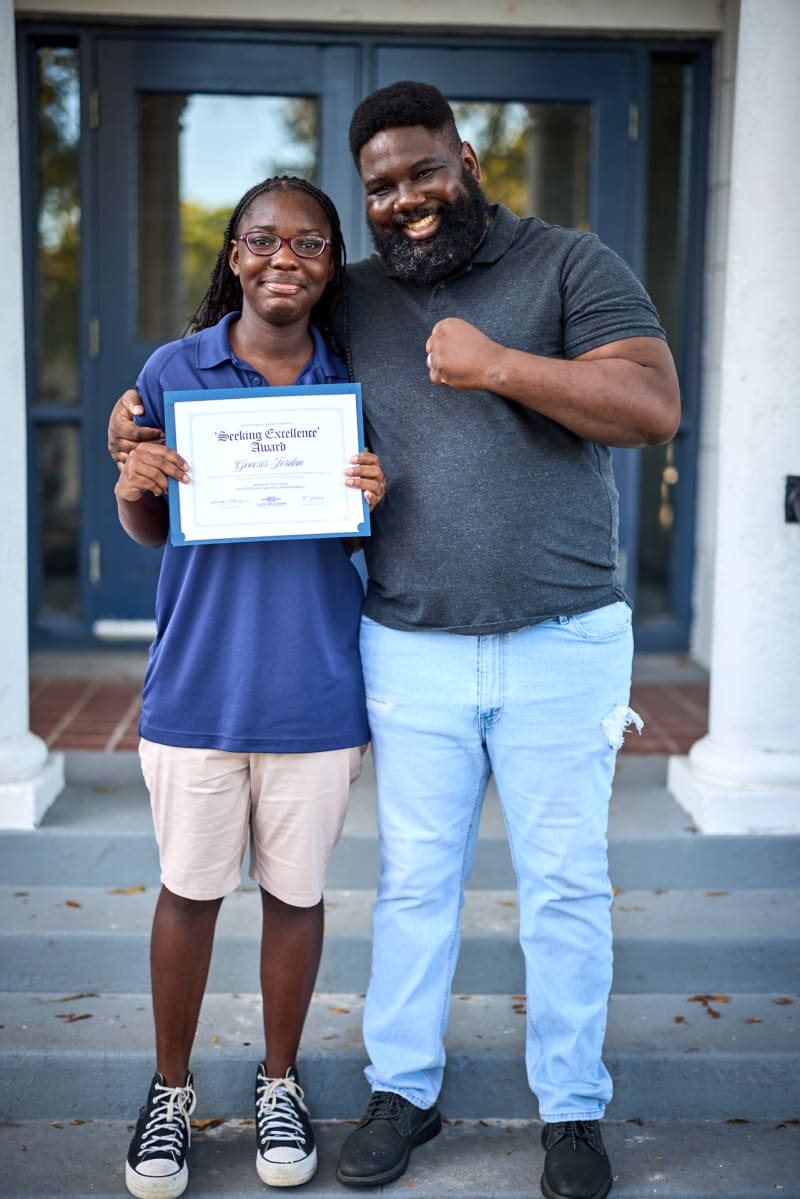 A student holding an award certificate beside a proud family member on the school steps