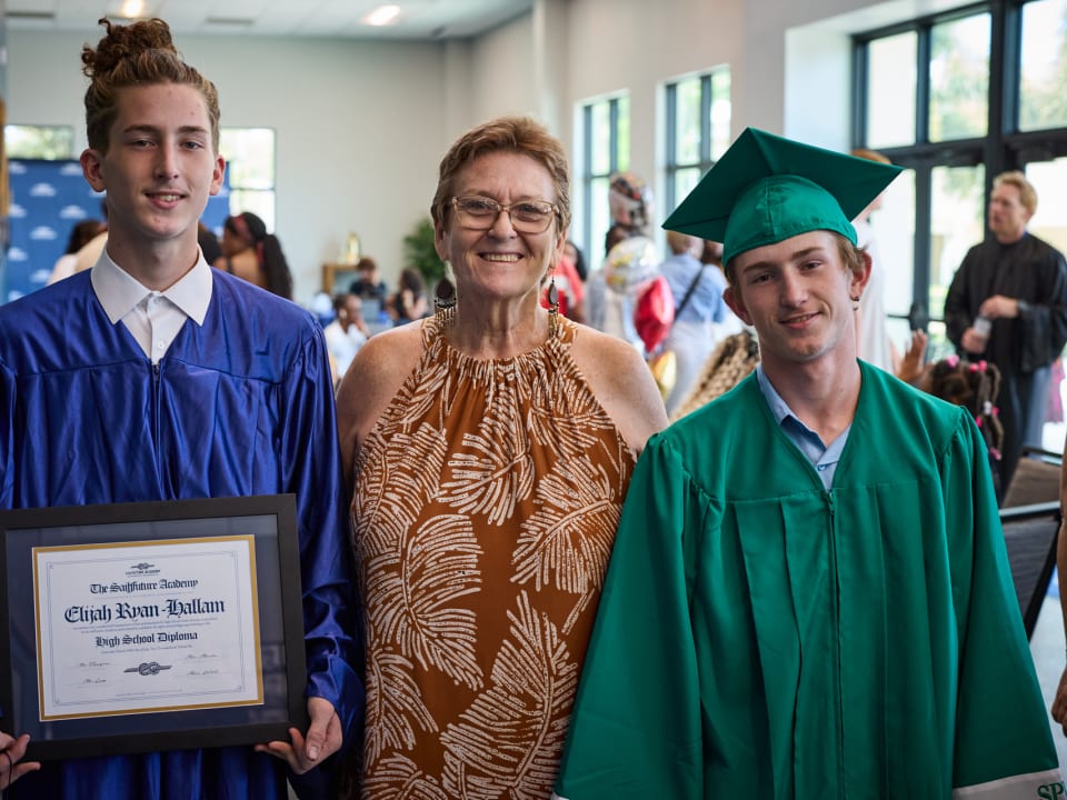 Two graduates pose with an adult mentor holding a diploma during the graduation ceremony.