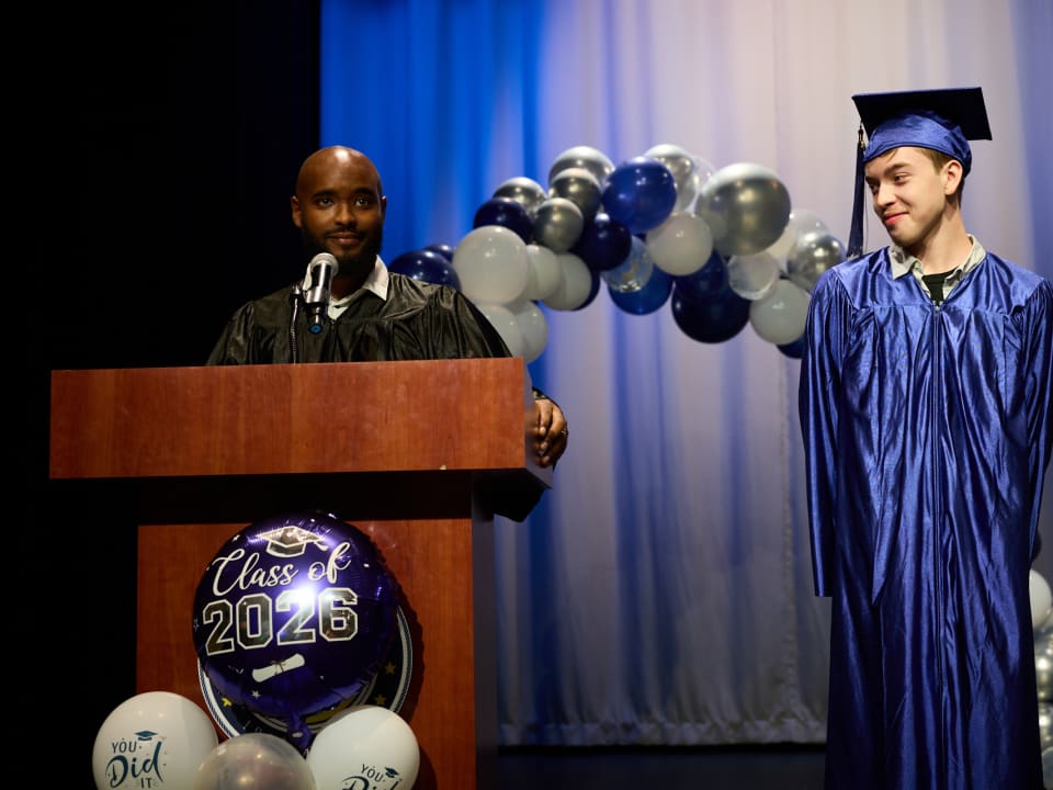 An instructor speaks at the podium while a student smiles during the graduation ceremony.