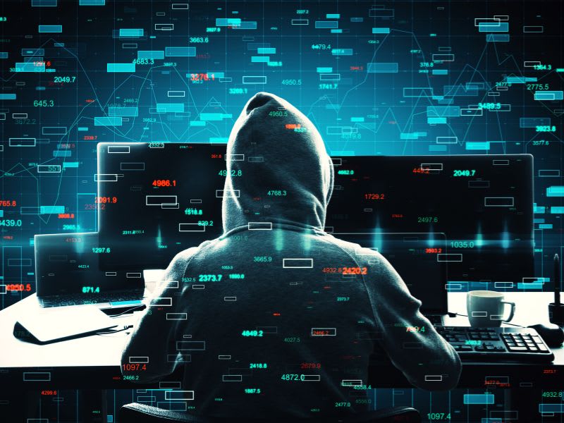 Hacker wearing a hoodie in front of multiple monitors with floating data graphics, symbolizing a malware or cyberattack