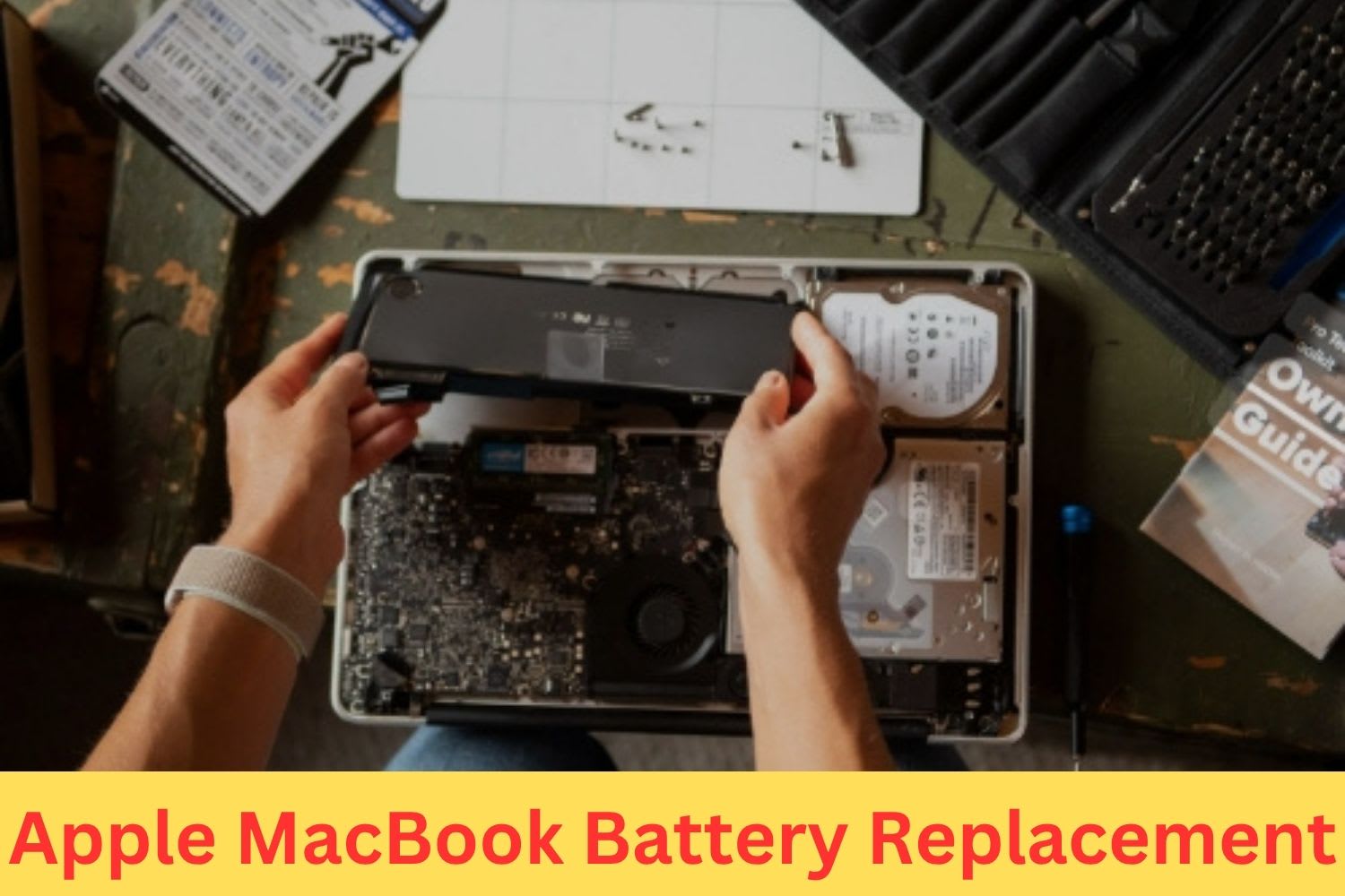 Technician replacing an Apple MacBook battery with the laptop opened on a desk, surrounded by repair tools and guidebooks