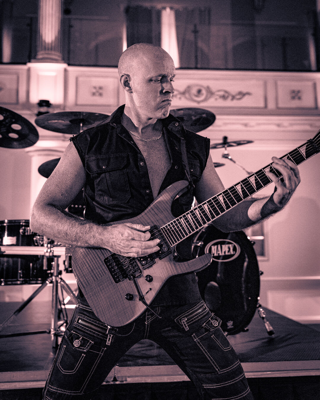 Le guitariste au crâne rasé et à la veste sans manches attaque un riff devant la batterie Mapex