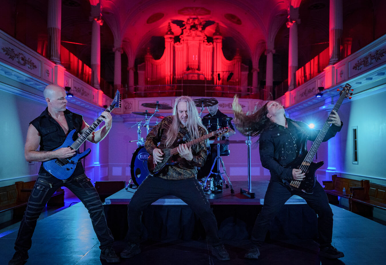 Trois musiciens de Forgotten Tales jouent côte à côte sur scène, cheveux au vent, devant l’orgue baigné de rouge