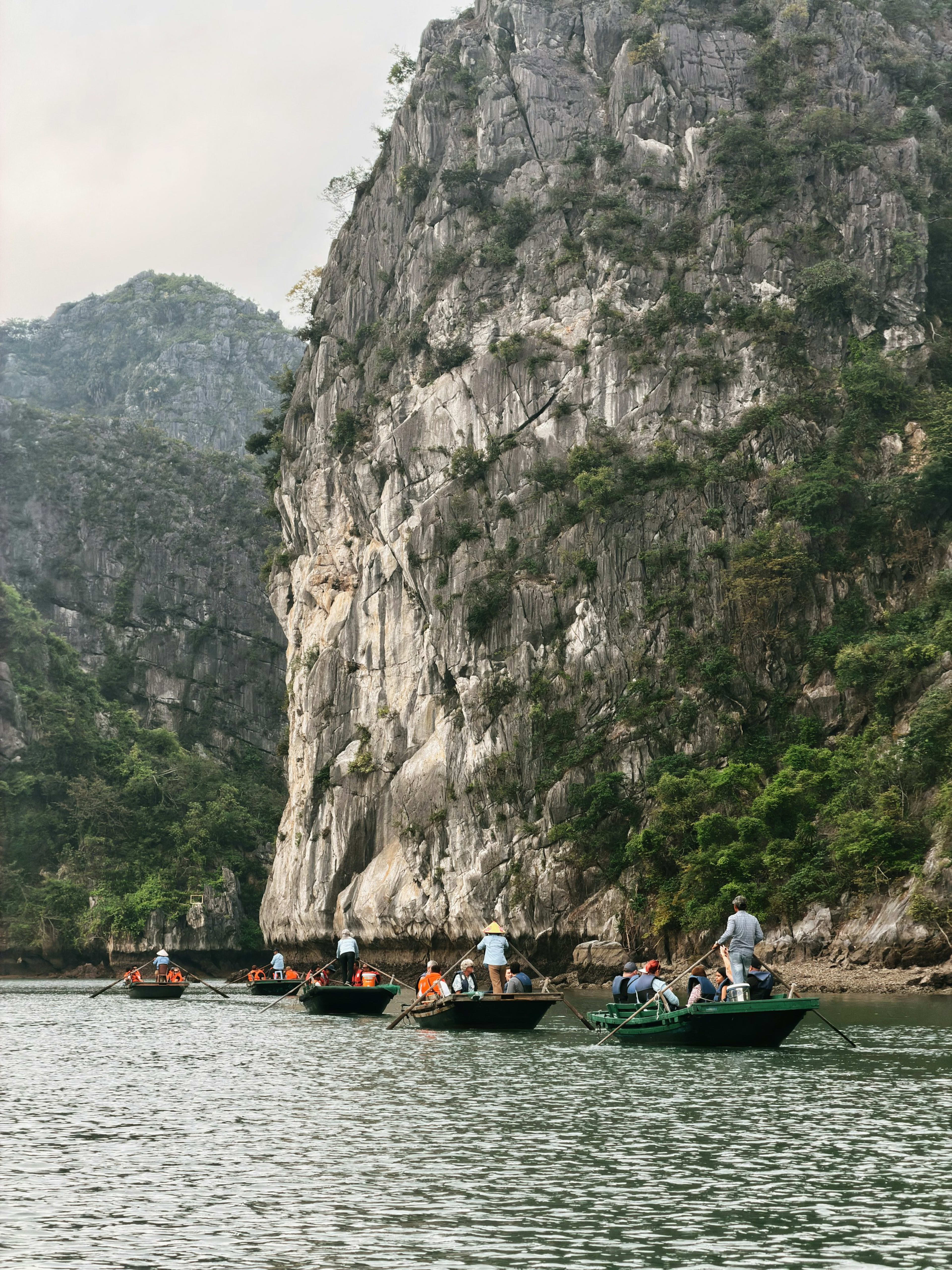 Rowing boats of visitors in orange life jackets gliding beneath a huge grey limestone cliff that drops straight into jade water.
