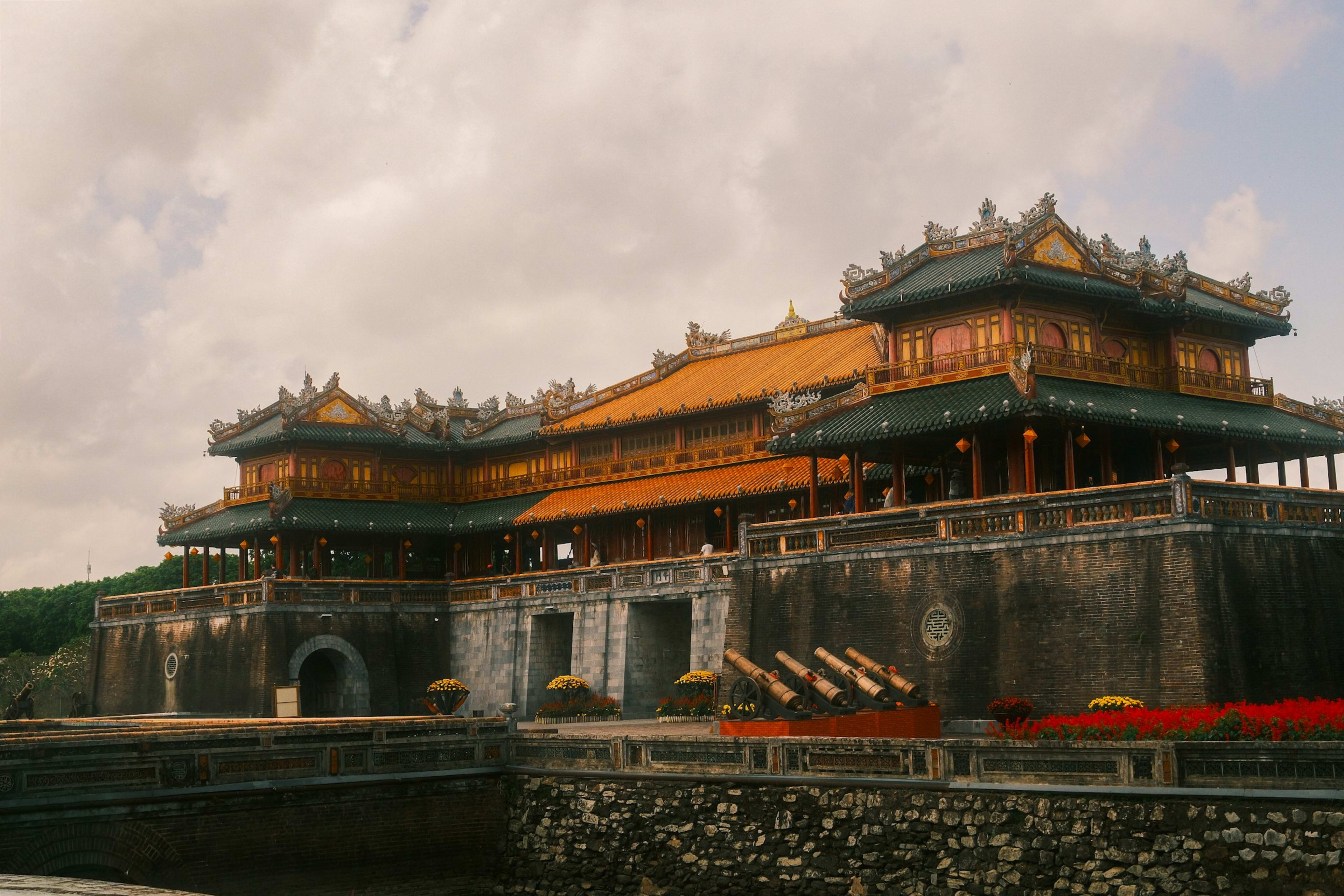 The tiered pavilion of the Huế citadel gate riding its stone rampart, glazed roof tiles catching the light and potted chrysanthemums lining the ramps below.