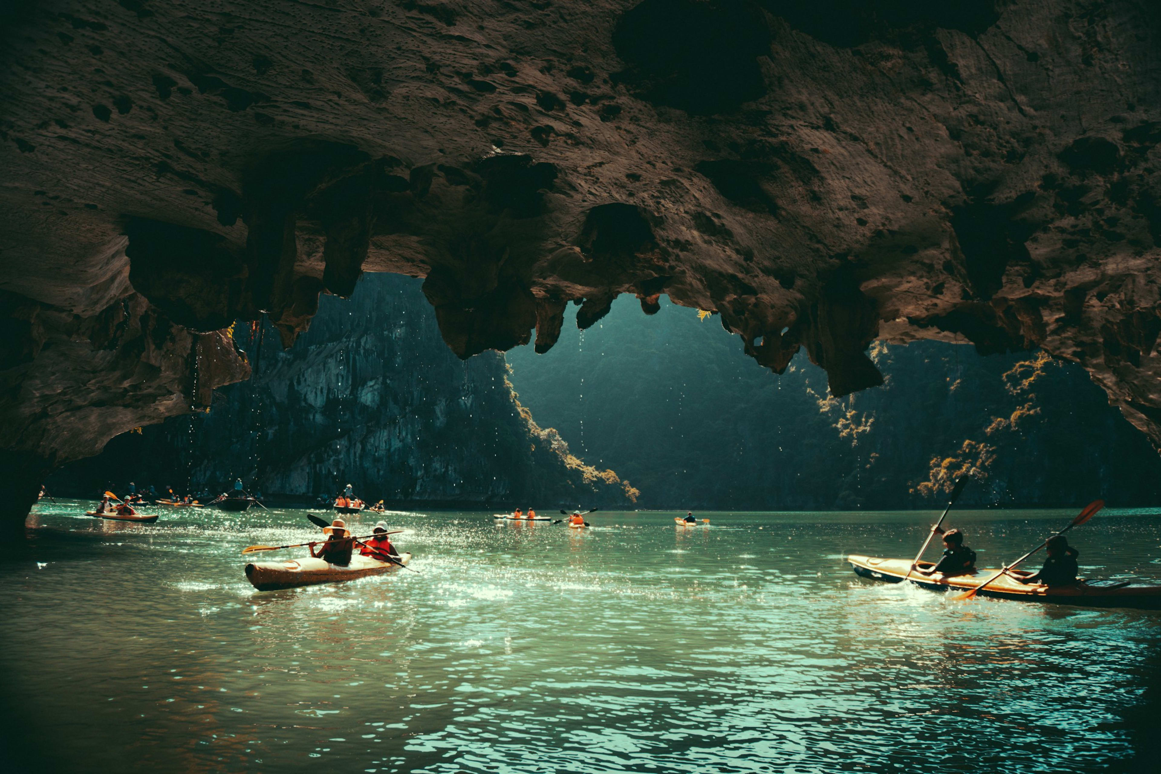 Kayakers paddling out through the mouth of a low sea cave, the rock ceiling framing daylight and a karst wall beyond.