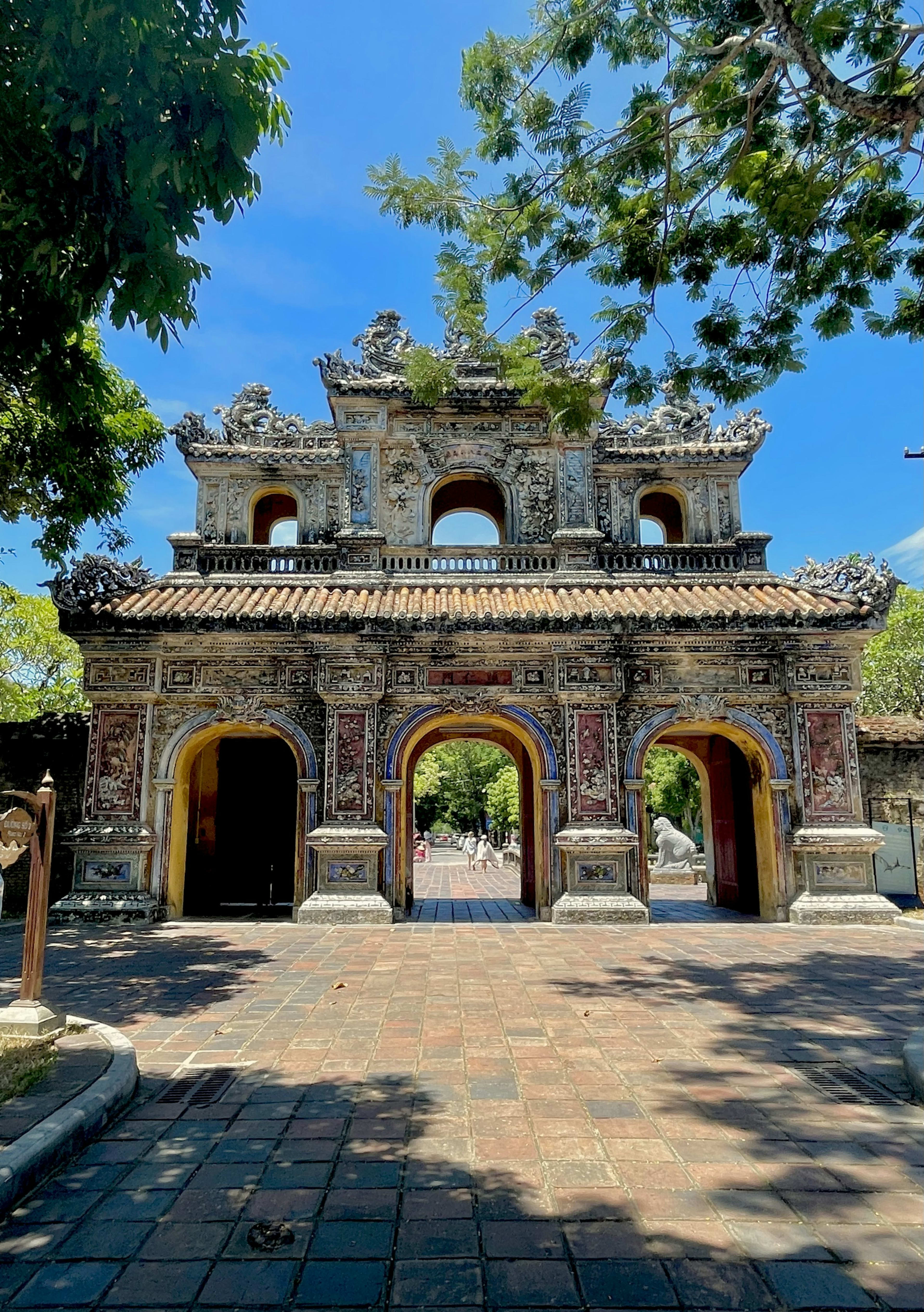 An ornate gate of the Huế citadel with three arched doorways, its porcelain-mosaic panels and tiled roof framed by overhanging branches.