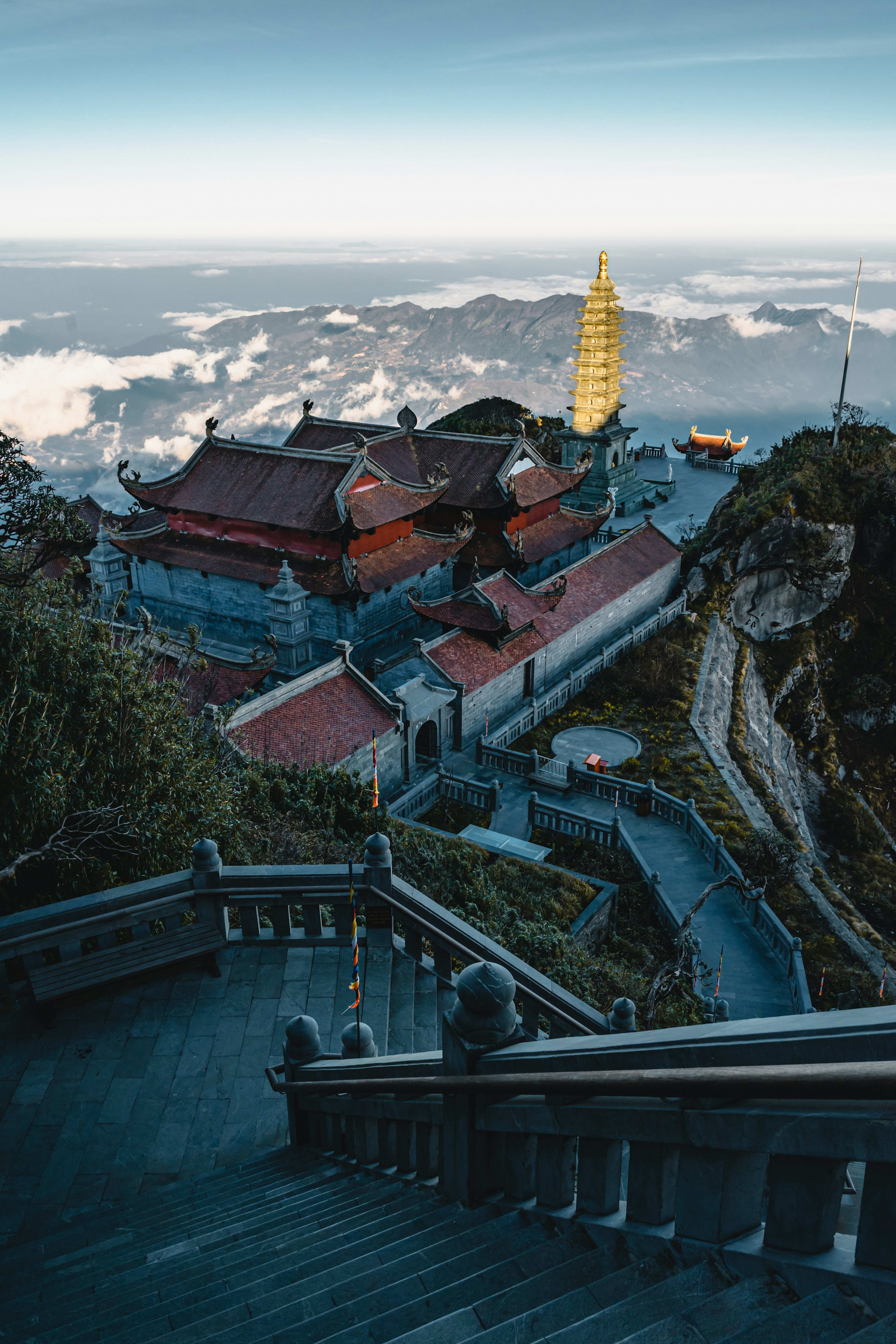 Tiered temple roofs and a golden pagoda standing above a sea of cloud at the summit of Fansipan, a broad stone stairway climbing toward them.