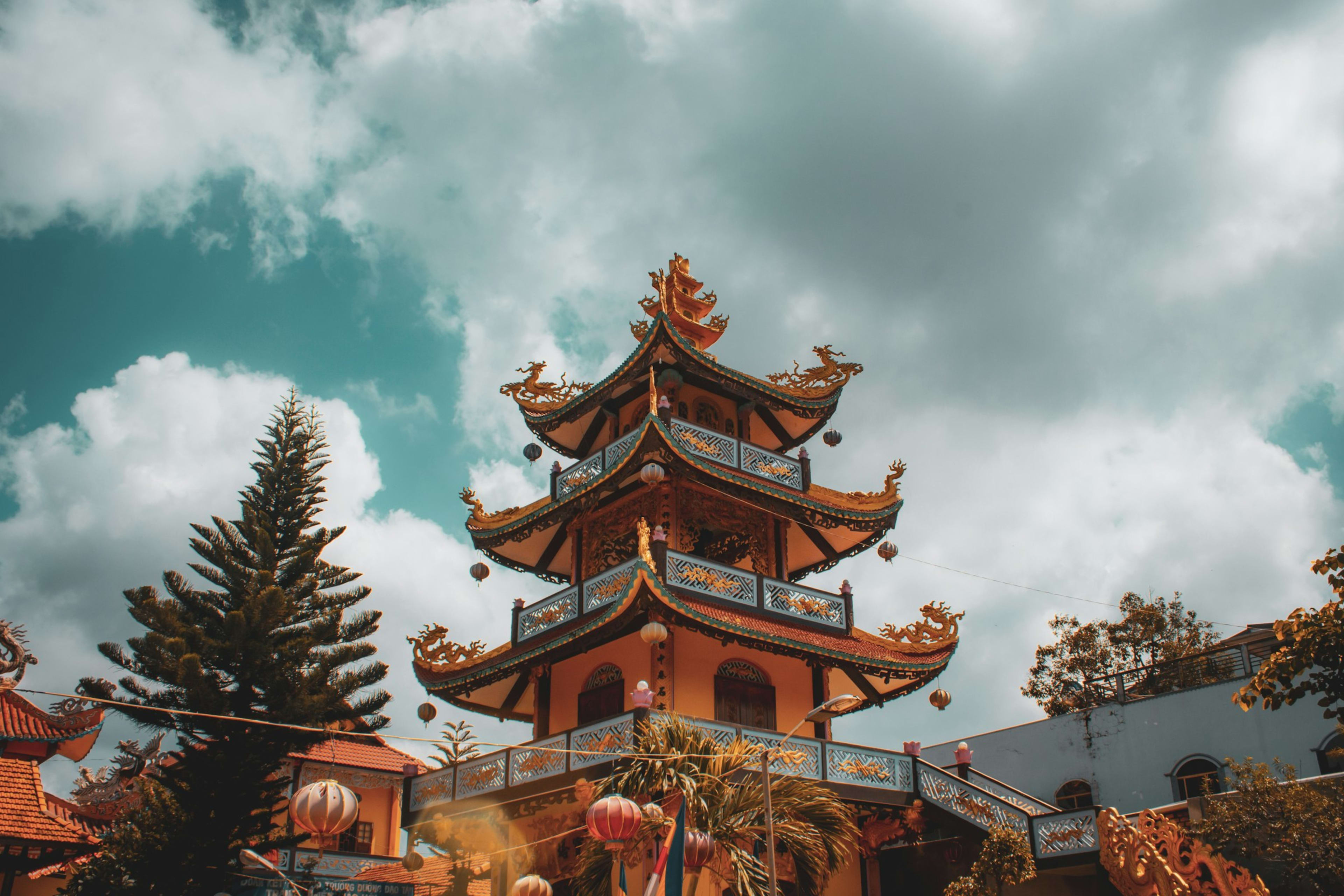 An ornate pagoda tower with gilded dragon-tipped eaves against a dramatic cloudy sky, a pine and red lanterns below it.