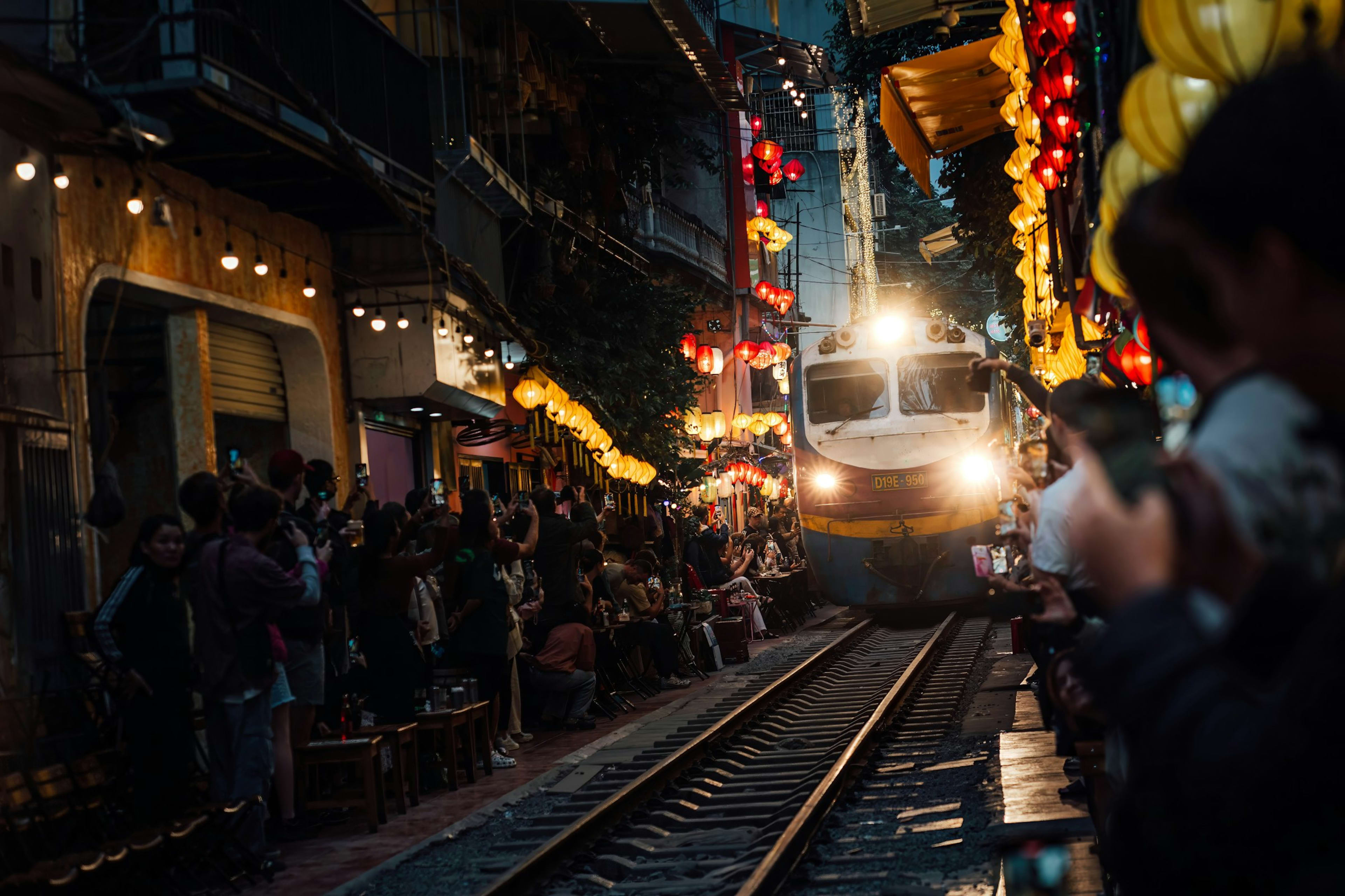 A train easing down the narrow lane of Hanoi Train Street at dusk, headlight on, while onlookers press back against café fronts hung with lanterns.
