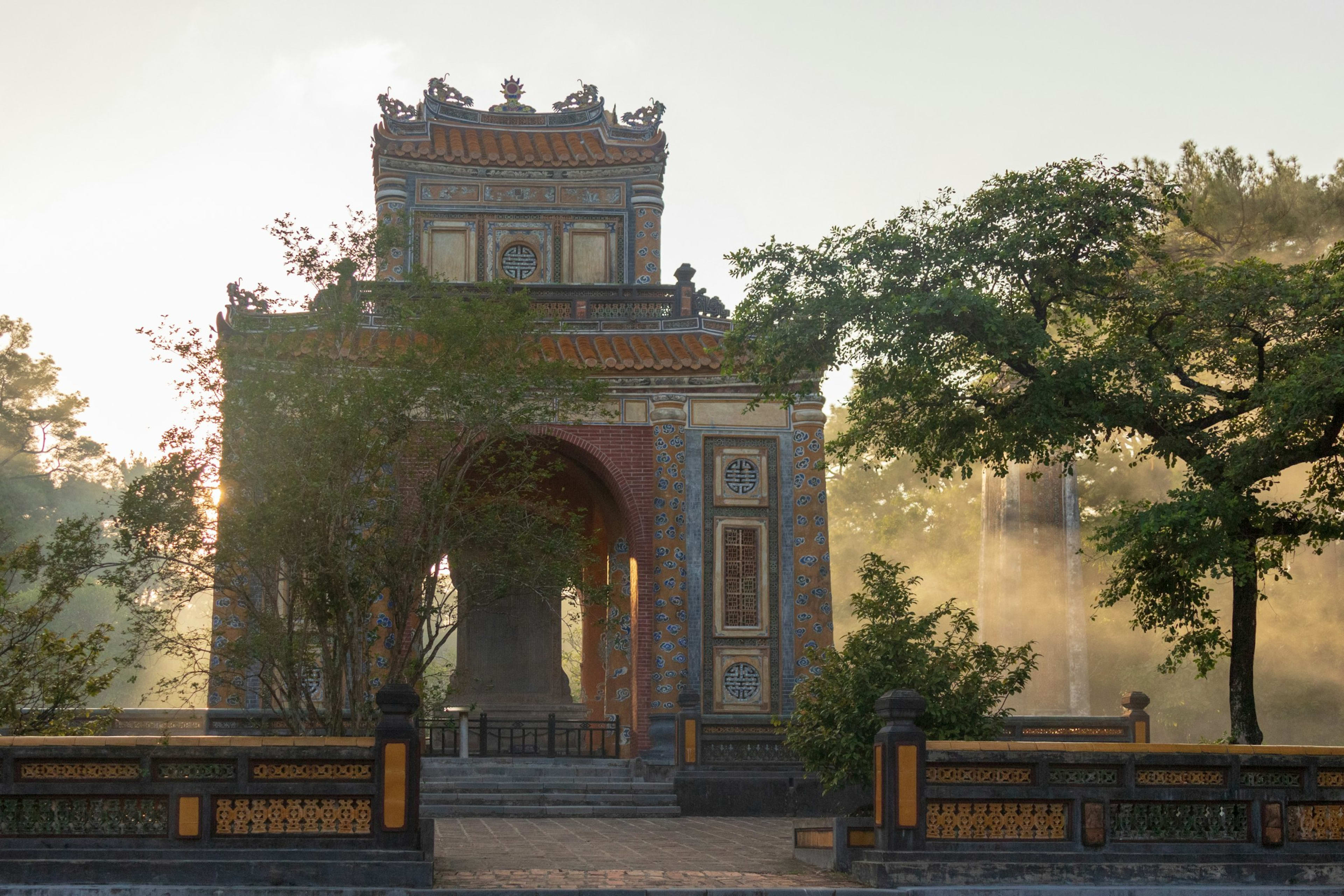 A stele pavilion at a Huế royal tomb catching low morning light through mist, its tiled roof and mosaic panels framing the stone tablet inside.