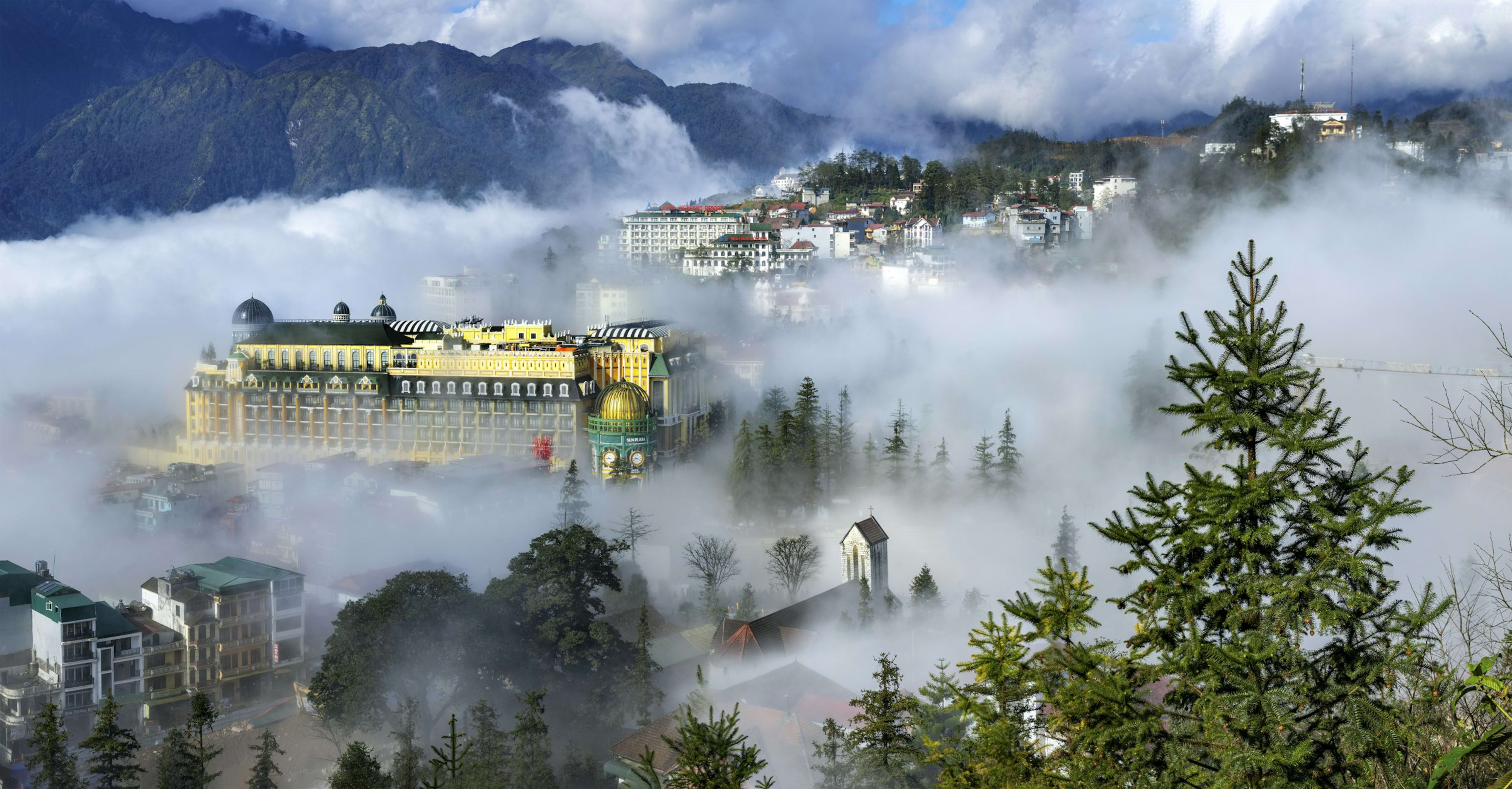 Sa Pa town half-swallowed by cloud, a hotel façade and pine tops rising out of the mist with mountains behind.
