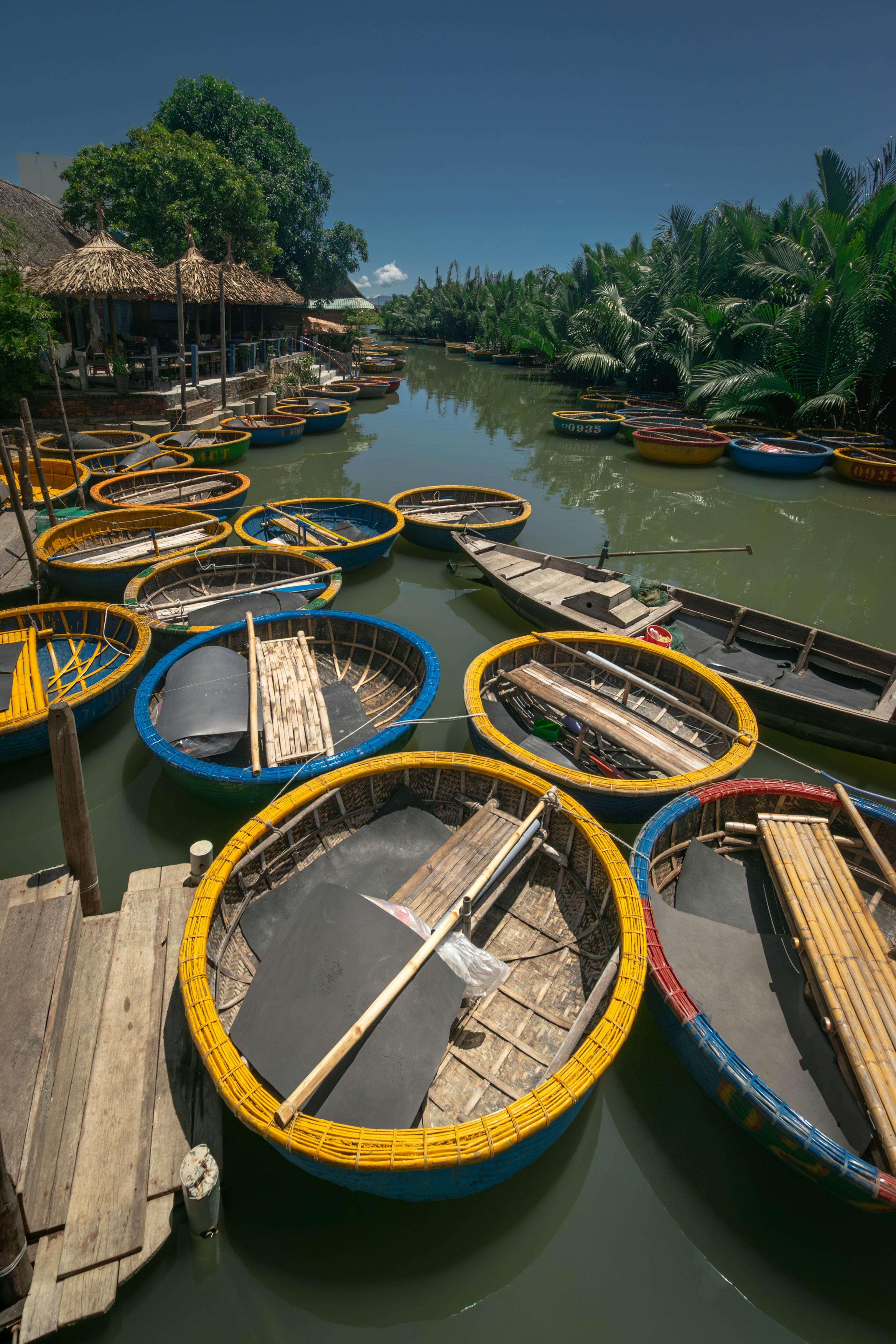 Round basket boats crowded along a river channel beside thatched huts, a bank of nipa palms on the far side.